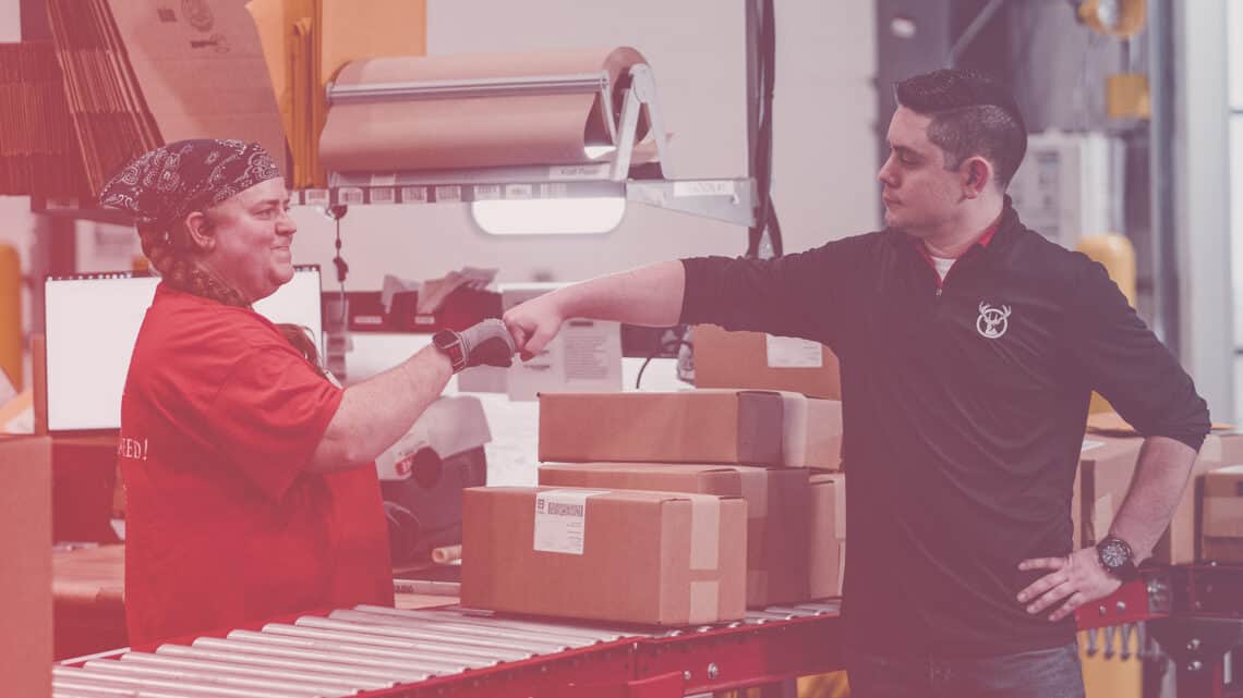 Two 3PL employees fist bump over a conveyer belt of packages